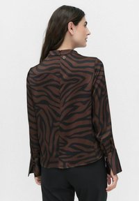 Blouse rayée marron et noire avec une texture lisse, col haut, et fermeture à boutons à l'arrière, dotée de manches longues et de détails aux poignets.