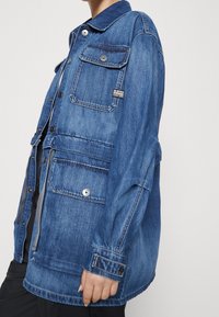 Veste en denim bleu moyen avec un devant boutonné, cinq poches et un col classique. Dotée d'une étiquette tissée sur le côté.