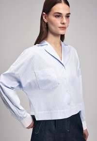 Camicia a maniche corte in blu chiaro e righe bianche, con taschino sul petto, maniche oversize e polsini bianchi a contrasto.