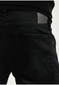 Jeans de mezclilla negra con un diseño ajustado, que presentan un parche de cuero en la cintura y un detalle distintivo de bolsillo trasero en ángulo.