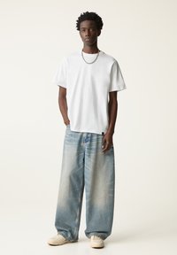Weißes Baumwoll-T-Shirt, kurze Ärmel, lockere Passform. Helle, baggy Jeans in Blau mit beschädigten Akzenten. Weißes Sneaker, silberne Kettenhalskette.