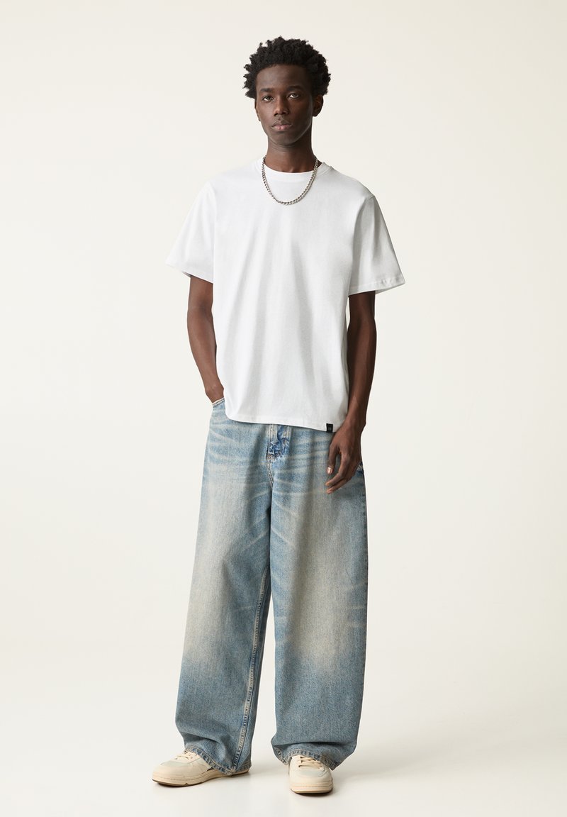 Weißes Baumwoll-T-Shirt, kurze Ärmel, lockere Passform. Helle, baggy Jeans in Blau mit beschädigten Akzenten. Weißes Sneaker, silberne Kettenhalskette.