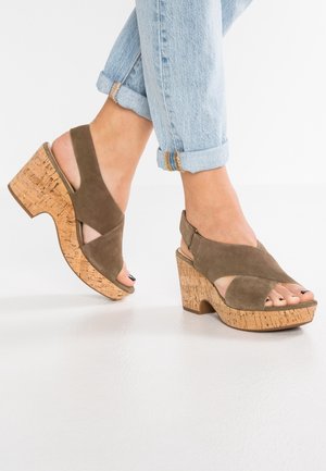 Pieds portant un jean bleu clair retroussé et des sandales compensées en liège en daim marron, avec des orteils ouverts et des brides arrière.
