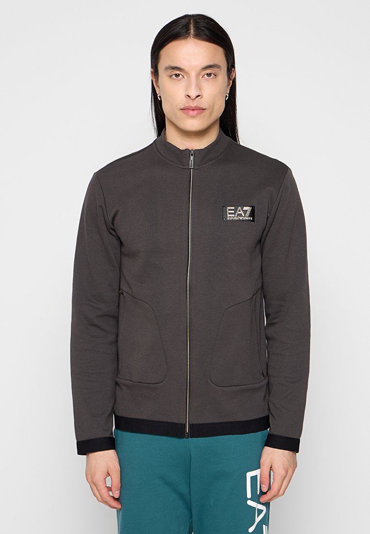 Giaccone zip in grigio scuro con colletto alto, polsini a coste e due tasche frontali. Presenta un patch con il logo sul petto sinistro. Texture liscia.