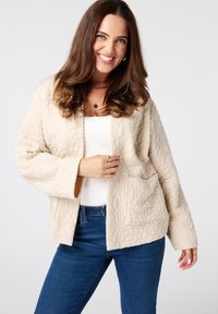 Beige gewatteerde jas met een gestructureerd bladerenpatroon, open voorkant, zijzakken en brede mouwen, gedragen over een witte top en blauwe spijkerbroek.