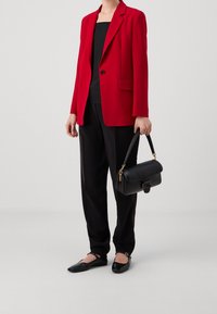 Červený blazer s klopami přes černý top, kombinovaný s černými kalhotami a černými lodičkami; držící malou černou kabelku se zlatými akcenty.