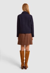 Giacca in misto lana blu navy con colletto a rever, abbinata a una gonna con motivo floreale marrone e stivali in suede beige al ginocchio.