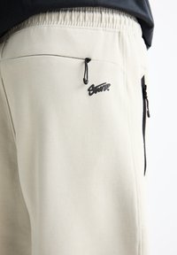 Helle beigefarbene Sweatpants aus weichem Material, mit einem schwarzen Logo, elastischem Bund und verstellbarem Kordelzug. Mit Reißverschlusstaschen.