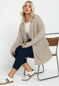 Lätt beige kofta med öppen front, ribbad mudd och texturerad tyg, parat med en vit t-shirt och mörk denimbyxor.