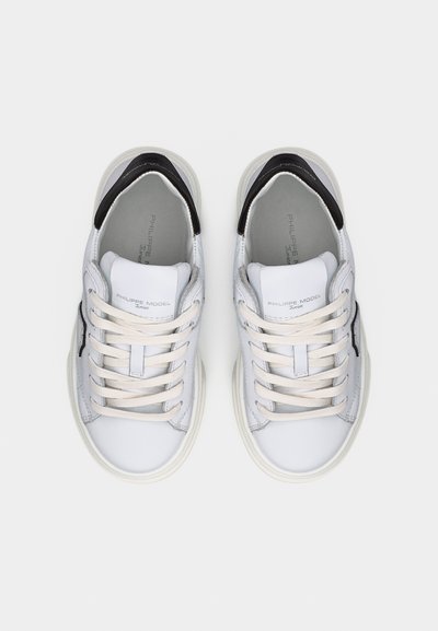 Paire de baskets basses blanches avec des accents noirs au talon et des lacets blancs, vue de dessus sur un fond uni.