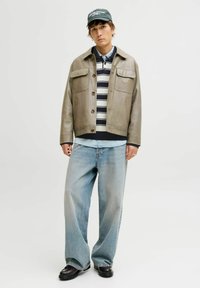 Šedá kožená bunda s dvěma předními kapsami, pod ní pruhovaná košile, modré džíny a černé boty. Model má na hlavě zelenou čepici s nápisem.