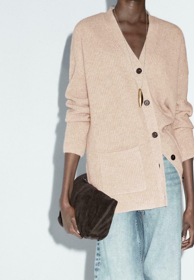 Une personne porte un cardigan en maille beige avec de gros boutons et un jean bleu clair, tenant une pochette pliante en daim marron foncé dans sa main gauche.