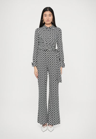 Tuta fantasia in bianco e nero con camicia a collo, maniche lunghe e pantaloni svasati, caratterizzata da un dettaglio in vita annodato e un design geometrico.