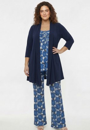 Kvinde med krøllet hår iført en blå top og bukser med blomsterprint samt en marineblå åben cardigan, står med den ene hånd på hoften.
