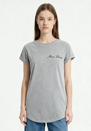 Jeune femme portant un t-shirt gris à manches retroussées avec le texte « Mira Paris » sur la poitrine et un jean bleu, debout devant un fond clair uni.