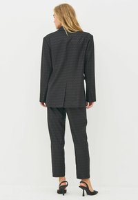 Woman standing with back to camera wearing a dark patterned suit with long blazer and fitted pants, and black high-heeled slingback shoes.