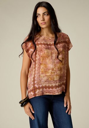 Femme aux longs cheveux foncés portant un chemisier à manches courtes à motif rose et marron, un jean bleu et des bracelets noirs à son poignet gauche.