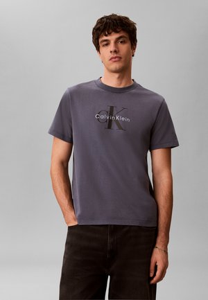 Młody mężczyzna z krótkimi kręconymi włosami, ubrany w szarą koszulkę Calvin Klein i czarne spodnie, stojący na jednolitym jasnoszarym tle.