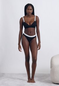 Fekete fehérnemű szett, amely egy vékony fehér pántos bralettből és egy fehér derékpántos, hozzáillő bikini alsóból áll, semleges háttér előtt.