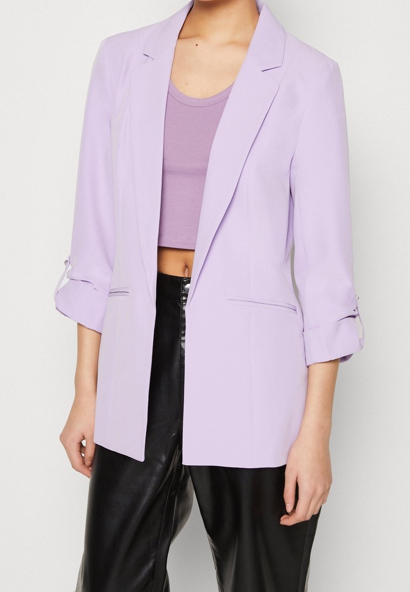 Blazer lilas à coupe ajustée, avec revers crantés et manches trois-quarts. Présente un design minimaliste et une texture lisse.