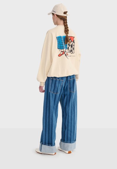 Person mit einem langen Zopf, der eine beige Mütze, einen beigen Pullover mit buntem Motiv auf der Rückseite, weite blau gestreifte Jeans und weiße Turnschuhe trägt.