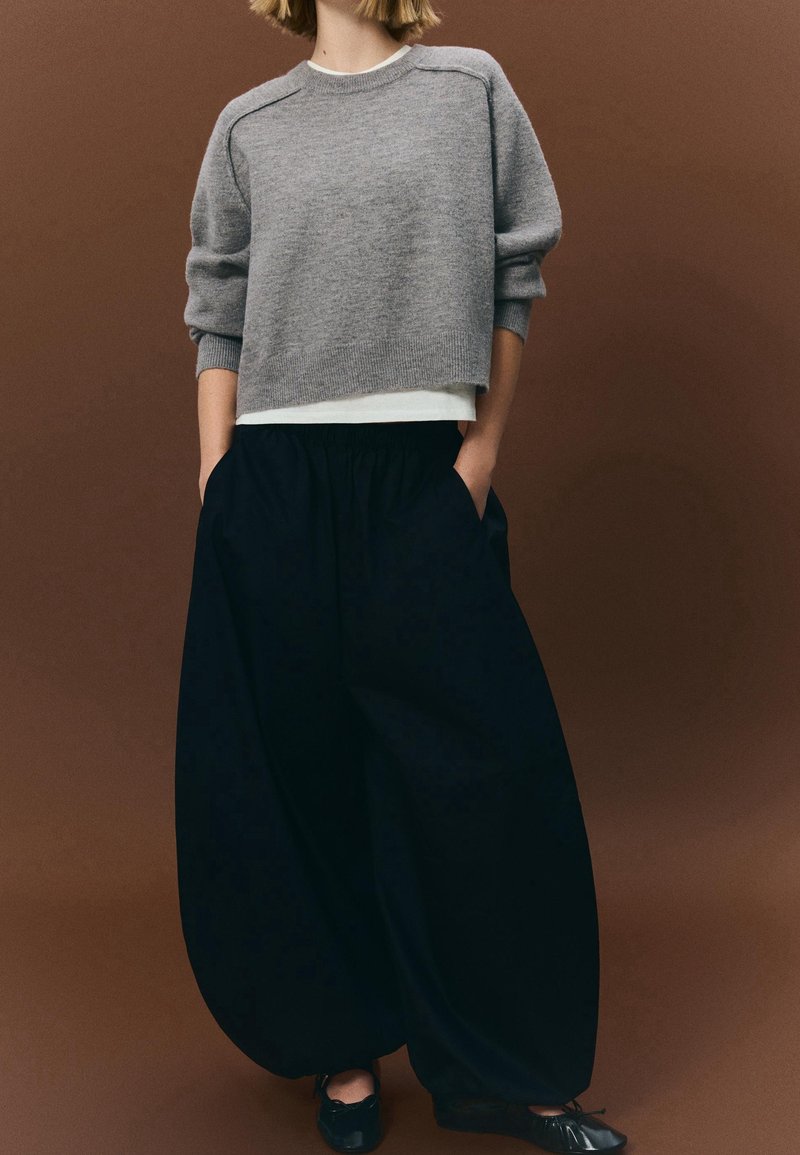 Personne portant un pull court gris, un sous-vêtement blanc, un pantalon large noir avec les mains dans les poches, et des chaussures noires sur un fond marron.