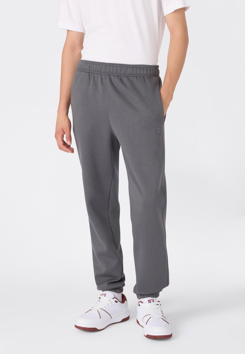Persoon in grijze joggingbroek met elastische boorden, witte sneakers met bruine accenten en een wit T-shirt, staand met één hand in de zak.