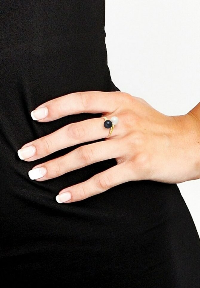Bague en or avec trois accents sphériques : un noir, un blanc et un plus petit en gris. Main avec des ongles soigneusement manucurés sur un fond noir.