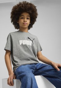 Pelēkas īsām piedurknēm t-krekls ar lielu balto "PUMA" logotipu un melnu pumus grafiku; kombinēts ar ziliem džinsiem.