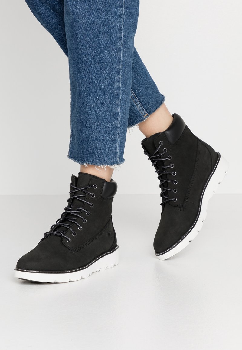 Bottines noires à lacets avec col rembourré et semelles blanches rainurées, portées avec un jean bleu court.