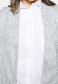 Šedý pletený cardigan s žebrovanou texturou nosený přes bílé košili s knoflíky, která má přední kapsu a kulatý výstřih.