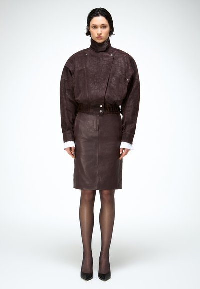 Žena u tamnobraon teksturiranom sako jaknom s visokim ovratnikom i pripadajućoj suknji do koljena, crne prozirne hulahopke i crne cipele s visokim potpeticama i špicastim vrhom.