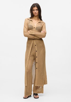 Femme portant un long cardigan beige ajouré et un pantalon large assorti, debout les bras croisés, chaussée de sandales noires ouvertes.