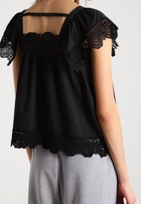 Blusa nera con dettagli in pizzo sulle maniche e sull'orlo, vestibilità rilassata, scollatura rotonda e apertura posteriore con dettaglio a cintura.