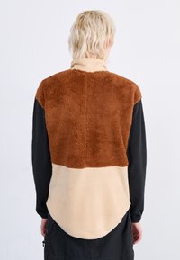 Chaqueta de forro polar marrón y beige con una parte superior texturizada y suave, y una parte inferior lisa. Presenta un dobladillo redondeado y un cuello alto.