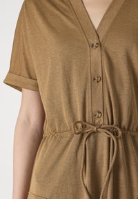 Îmbrăcăminte din material maro cu decolteu în V și mâneci scurte răsucite. Dispune de trei nasturi, un cordon la talie și buzunare frontale.