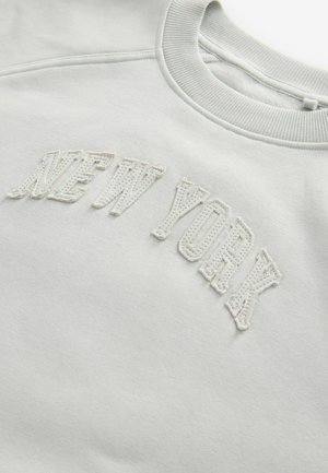 Ljusgrå sweatshirt med ribbat halsband. Framsidan har vit broderad text "NEW YORK" i en fet, texturerad stil. Mjuk tygkvalitet.