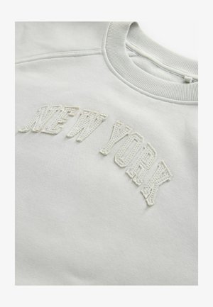 Vaalean harmaa collegepaita, jossa on ribbauskaulus. Etupuolella on valkoista brodeerattua tekstiä "NEW YORK" rohkealla, teksturoidulla fontilla. Pehmeä kangas.