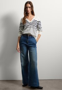 Donna che indossa un maglione bianco con righe blu scuro, jeans a gamba larga blu, scarpe nere, in piedi contro un muro bianco semplice, con una mano in tasca.