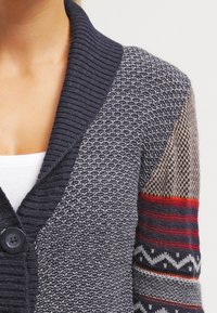 Sweter wykonany na drutach z głębokim granatowym szalowym kołnierzem, teksturowanym ciałem i wzorzystymi rękawami z czerwonymi, beżowymi i niebieskimi akcentami. Dwie widoczne guziki.