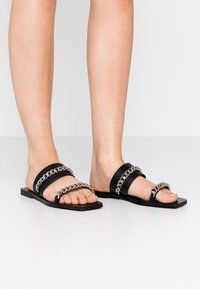 Svarta platta sandaler med breda remmar och silverkedjeaccenter, med en slät texturerad sula och minimalistisk design.