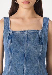 Rochie din denim cu decolteu pătrat, bretele groase și detalii plisate. Materialul este de un albastru mediu, cu linii de cusătură vizibile.