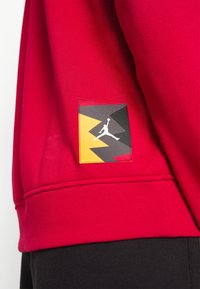 Sudadera roja con un tejido texturizado. La manga izquierda presenta un parche con un diseño geométrico dinámico en negro, amarillo y rojo con un logotipo.