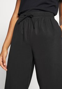 Personne portant un pantalon noir ample avec une taille élastique et un cordon de serrage noué, se tenant avec une main dans la poche et l'autre détendue.