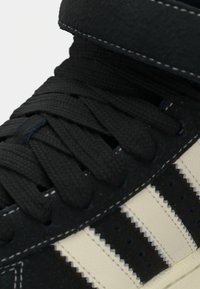 Μαύρο αθλητικό παπούτσι με λείο σουέντ φινίρισμα, υφασμένα κορδόνια και κρεμ ρίγες. Υψηλή σχεδίαση με επενδεδυμένο γιακά και άσπρη ραφή.