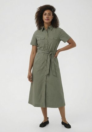 Žena s kučeravými vlasmi, oblečená v zapínaných midi šatách olivovej farby s opaskom v páse a čiernych plochých topánkach, stojaca na bielom pozadí.