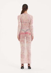 Průhledné růžové šaty dlouhého rukávu s květinovým vzorem, těsný střih a detail loga na zádech. Nosí se s vysokými podpatky.