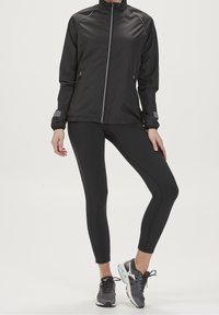 Chaqueta negra ligera con cuello alto y cierre de cremallera, combinada con leggings negros. Detalles texturizados en las mangas, llevada con zapatillas de correr grises.