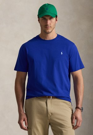 Mann trägt grüne Kappe, blaues T-Shirt mit Logo, beige Hose und Armbanduhr und steht vor einem einfarbigen Hintergrund.