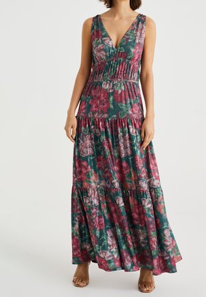 Femme portant une robe maxi sans manches à col en V profond avec des fleurs vertes, roses et rouges, associée à des sandales marron à bout ouvert.